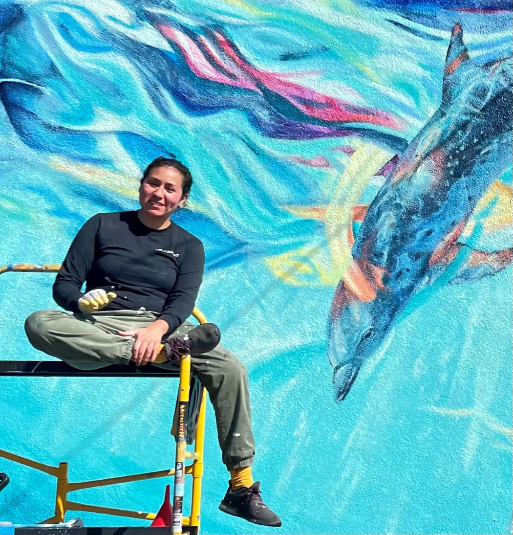 muralist,Myisha Arellano, sitting on scafolding, backdrop camous legacy mural