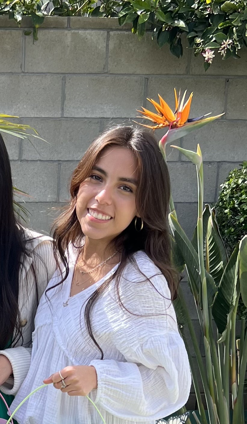 Isabela has her brown hair parted at the middle, is wearing gold hoop earings, and is wearing a white long sleeved blouse. She is smiling at the camera
