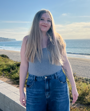 Taylor has blonde long hair. She is wearing a grey tank top and blue jeans. She is smiling directly at the camera.